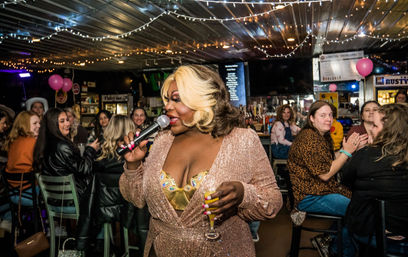 Drag performer in a sparkling rose-gold gown singing into a microphone and holding a cocktail, entertaining a seated crowd of women at a lively neighborhood bar decorated with string lights and pink balloons — vibrant nightlife drag show scene.