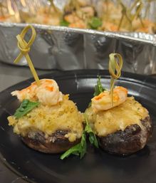 Two bite-sized stuffed mushroom appetizers topped with garlic-herb breadcrumb crust and mini shrimp skewers on a black plate, aluminum tray blurred in the background