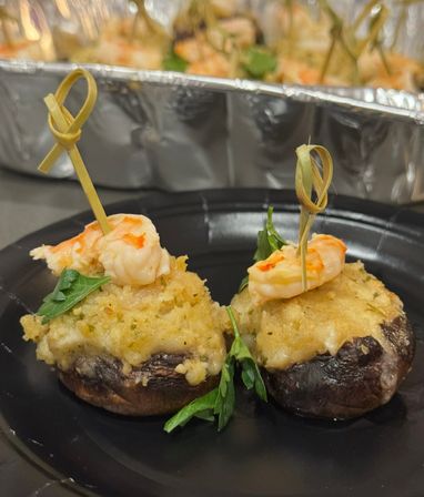 Two bite-sized stuffed mushroom appetizers topped with garlic-herb breadcrumb crust and mini shrimp skewers on a black plate, aluminum tray blurred in the background