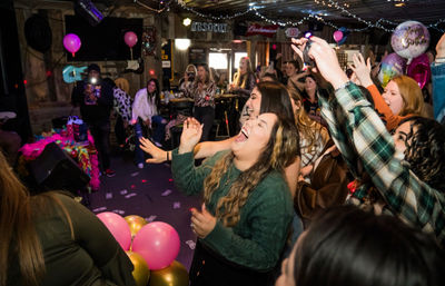 Lively indoor celebration at a bar-style venue: a crowd of people cheering and laughing amid pink and gold balloons, string lights and birthday decorations.
