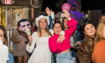 Lively bachelorette-style party at a bar: woman in white dress and cowboy hat holding cash and a face cutout while friends cheer and dance.