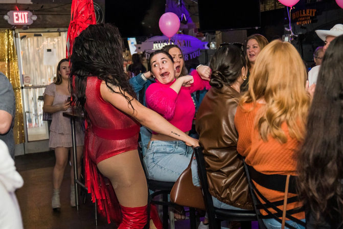 Nightlife bar scene with a performer in a red sequined bodysuit and thigh‑high boots entertaining a group of seated women; one woman in a bright pink sweater reacts with a surprised face as pink balloons float overhead.
