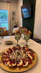 Close-up of a Caprese platter with burrata and balsamic on a kitchen island, surrounded by individual charcuterie-and-cheese cups in green goblets, a pesto bowl, and a modern living room background.