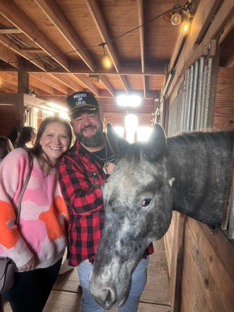 Smiling couple in a rustic wooden barn posing with a gray horse leaning out of a stall, string lights overhead, man in red plaid shirt and cap and woman in a pink sweater.