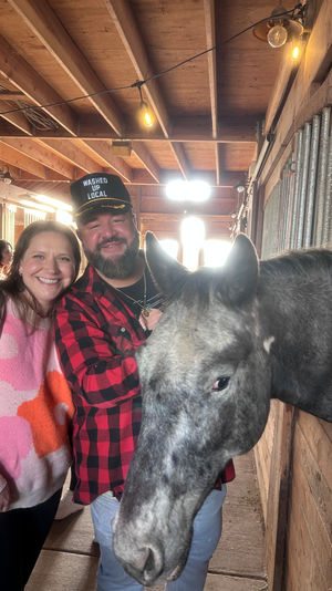 Smiling couple in a rustic wooden barn posing with a gray horse leaning out of a stall, string lights overhead, man in red plaid shirt and cap and woman in a pink sweater.