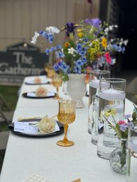 Cheerful outdoor garden table setting on a white tablecloth with an amber vintage goblet, black plates topped with pale roses, tall glass cylinders holding floating candles, and a colorful wildflower centerpiece.