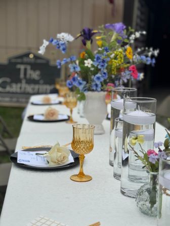 Cheerful outdoor garden table setting on a white tablecloth with an amber vintage goblet, black plates topped with pale roses, tall glass cylinders holding floating candles, and a colorful wildflower centerpiece.