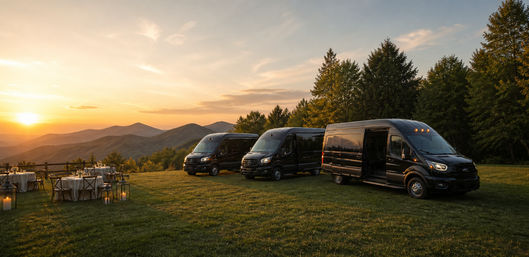 Three sleek black passenger vans parked on a grassy mountain overlook at sunset beside candlelit outdoor dining tables and lanterns.
