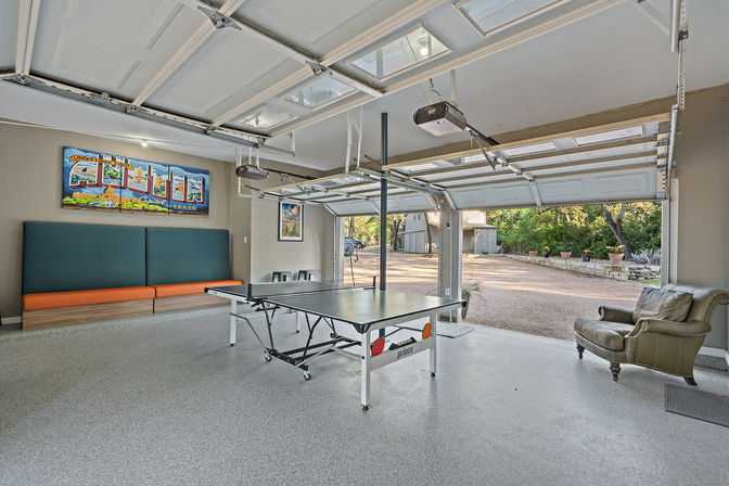 Bright Austin, Texas garage game room with ping-pong table, teal-and-orange bench seating, leather armchair, colorful Austin wall art, and open garage doors overlooking a gravel driveway and trees.