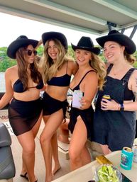Four smiling women in black bikinis and cowboy hats on a pontoon boat at a sunny lake, holding drinks and posing for a summer friends' boat party.