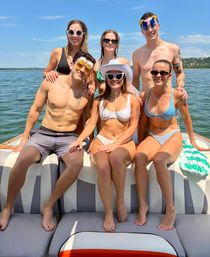 Six friends in swimwear posing and smiling on a motorboat at a sunny lake with clear blue sky and distant shoreline, some wearing novelty sunglasses and a white sun hat.