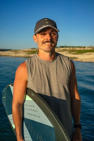 Smiling man in a baseball cap and gray sleeveless shirt holding a striped water-sport board by a sunlit lake shoreline under a clear blue sky.