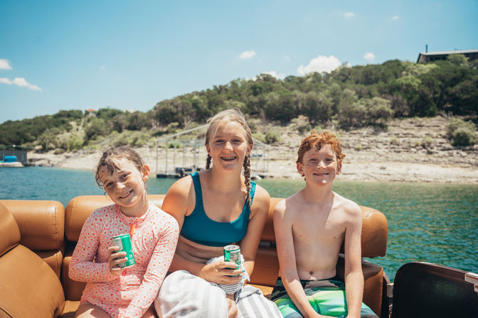 Three smiling kids in swimsuits sitting on a tan boat bench with canned drinks on a sunny lake, rocky shoreline, dock, and wooded hill in the background