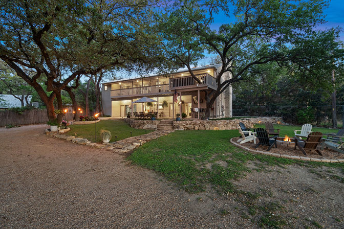 Dusk view of a two-story house with lit wraparound balcony, oak‑shaded backyard patio, stone terracing, gravel driveway and a fire pit ringed by Adirondack chairs.