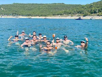 Group of friends floating in a clear blue lake near a wooded shoreline, holding canned drinks on a sunny summer day with boats in the distance.