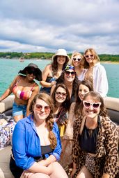 Group of women enjoying a summer lake boat ride, smiling in swimsuits, hats and heart-shaped sunglasses with green shoreline in the background