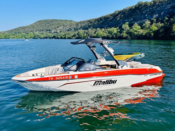Red-and-white wakeboard boat floating on a calm Texas lake, sunny day with forested cliffs and a wakeboard on the tower.