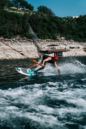 Action shot of a wakesurfer in a red cap crouching on a teal board, gripping a tow rope and carving spray across a lake with limestone cliffs and a boathouse in the background.
