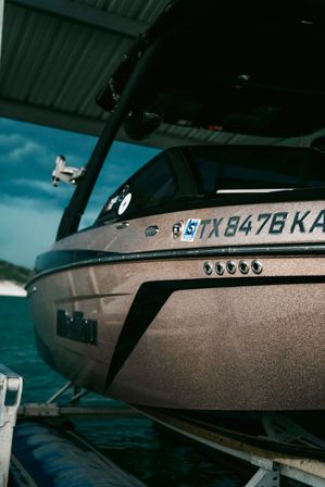Close-up of a metallic bronze wakeboard boat with Texas registration (TX) perched on a covered lift over turquoise lake water, angled bow and wakeboard tower visible.