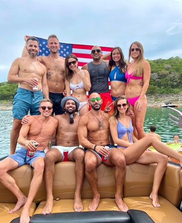Group of friends in swimwear posing on a boat with a large American flag, holding drinks on a summer lake with green, tree-lined rocky shoreline