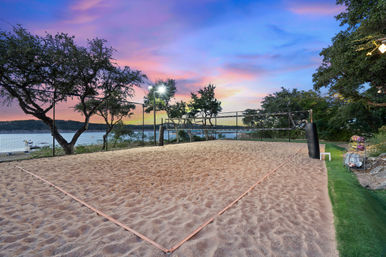 Lakeside sand volleyball court at sunset with net and lights framed by trees, colorful pink-blue sky and boats on the water beyond