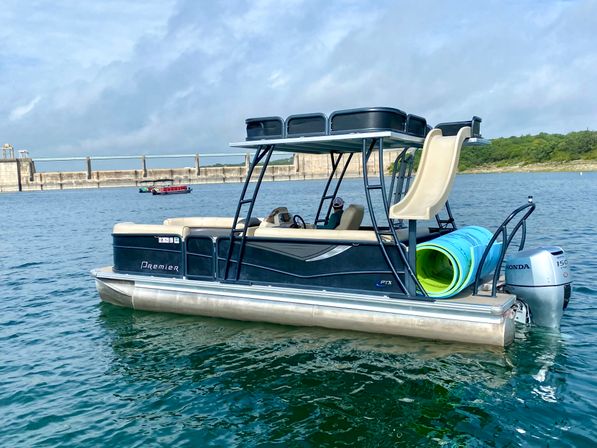 Pontoon boat with rooftop canopy and tan slide, colorful rolled-up water mats on the stern, floating on a blue lake near a concrete dam and tree-lined shoreline under a cloudy sky.