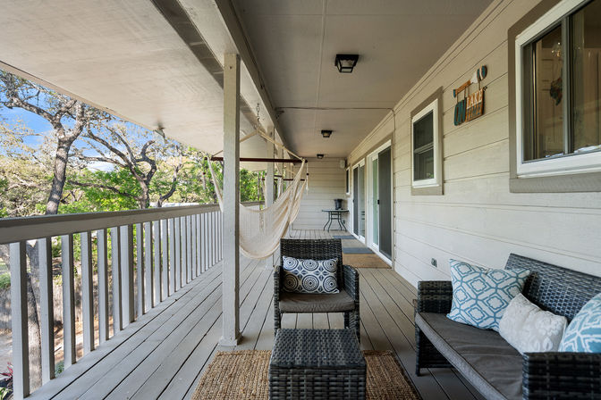 Sunny covered wooden deck with hanging hammock, wicker loveseat and chair with patterned cushions and jute rug, white railing and sliding doors overlooking leafy oak trees