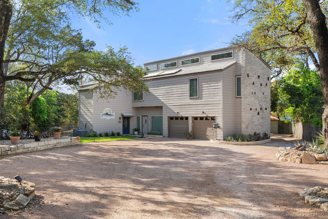 Sunlit contemporary two-story light-gray house with metal roof and clerestory windows, double garage and wide gravel driveway framed by oak trees, stone planters and potted succulents.