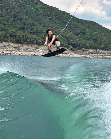 Wakeboarder soaring above a turquoise lake wake, clutching the tow handle as a boat-made wave curls toward a rocky shoreline and tree-covered green hills.