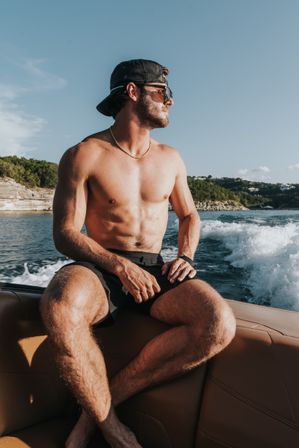 Shirtless man in a backward cap and sunglasses on a speedboat, sunlit torso and gold chain, cruising past a rocky green coastline with white wake on a blue summer sea