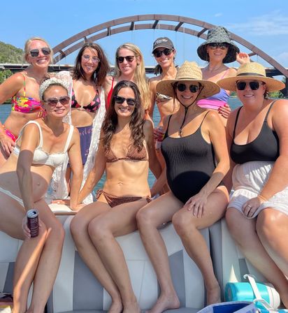 Group of friends on a boat under an arched bridge, smiling in swimsuits, sunglasses and sun hats on a sunny summer river cruise.
