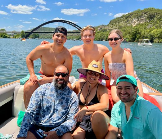Six friends smiling on a motorboat on Lake Austin with the Pennybacker (360) Bridge and sunny blue sky in the background — summer boating in Austin, Texas.