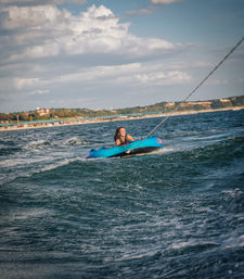Smiling woman in a life jacket tubing on a bright blue inflatable towed behind a boat across choppy lake water with shoreline and cloudy sky in the background