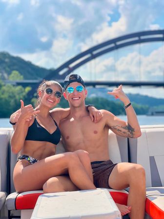 Smiling young couple in swimwear on a boat, flashing shaka signs and wearing mirrored sunglasses, arched bridge and tree-lined river in the background on a sunny summer day.