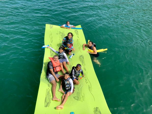 Family and kids smiling on a bright lime-green floating mat in clear green lake water, wearing life jackets and using swim noodles and water toys for a fun summer water activity.