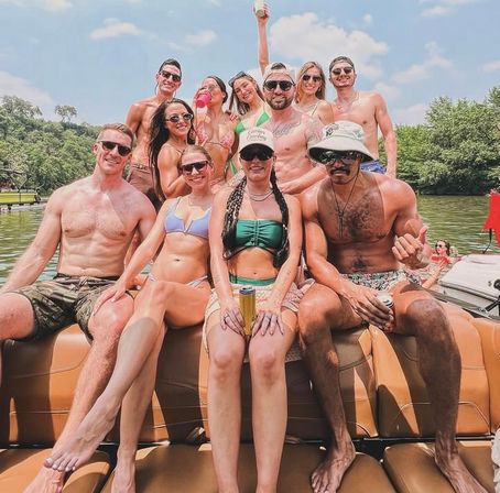 Group of friends in swimsuits enjoying a sunny boat party on a tree-lined lake, seated on tan boat seats and holding drinks.