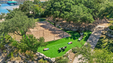 Aerial view of a sunny lakeside park with a sand volleyball court beside bright green artificial turf, cornhole boards and lounge chairs under oak trees, stone edging and a nearby dock.