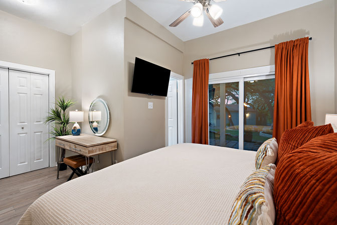 Cozy neutral bedroom with large bed, rust-orange pillows and curtains, sliding glass door to backyard, wall-mounted TV, rustic desk with round mirror and lamp