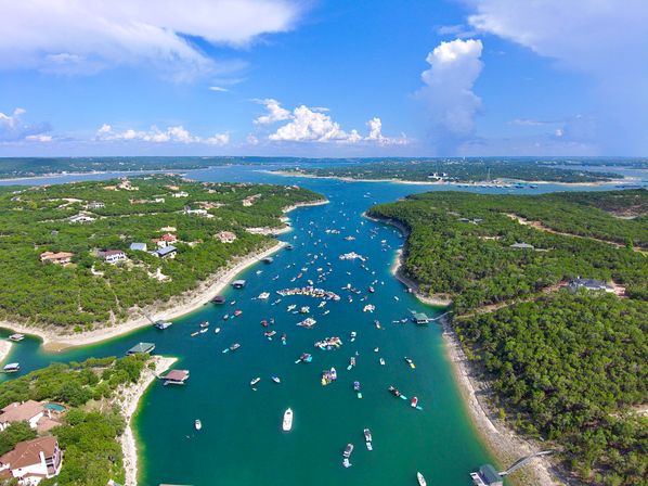 Aerial view of a turquoise lake channel filled with dozens of boats and floating rafts, flanked by green tree‑covered shorelines and docks under a bright blue sky with puffy clouds.