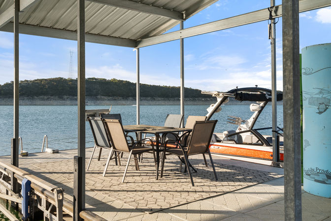 Covered lake dock with outdoor dining table and six chairs beside a parked wakeboard boat in a boat slip, calm blue water and wooded shoreline under a sunny sky.