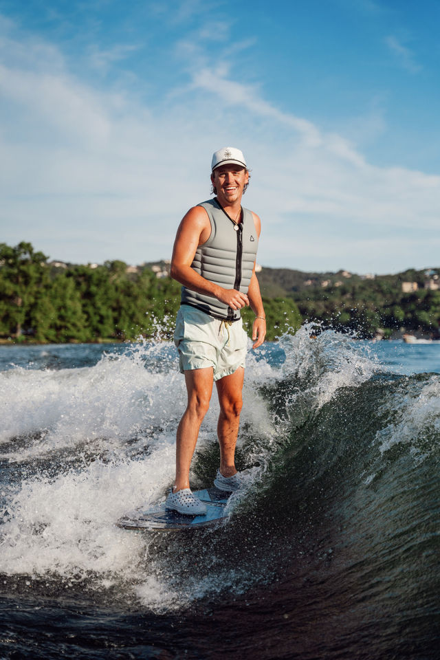 Smiling man in a gray life vest and white cap wakesurfing on a sunny lake, riding a wake with wooded shoreline and blue sky