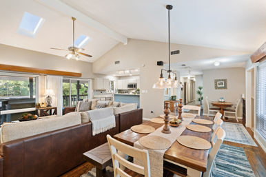 Sunlit open-concept living and dining area with vaulted ceiling and skylights, leather sectional sofa, wooden dining table set with woven placemats under pendant lights, white kitchen and sliding glass doors to a tree-lined deck.