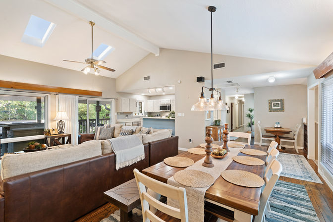 Sunlit open-concept living and dining area with vaulted ceiling and skylights, leather sectional sofa, wooden dining table set with woven placemats under pendant lights, white kitchen and sliding glass doors to a tree-lined deck.
