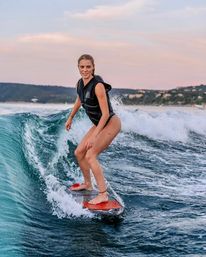 Young woman wakesurfing on an orange board across a turquoise wave at sunset with a distant coastline in the background