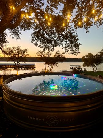 Lakefront sunset with warm string lights strung through oak branches, a round metal stock-tank pool in the foreground reflecting multicolored floating LED lights, calm lake and boat docks in the background.