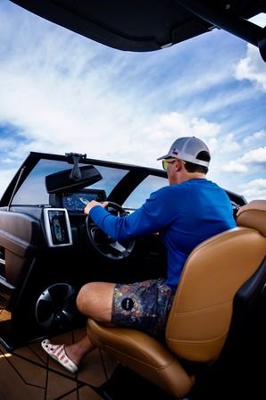Person steering a motorboat at the helm, using a digital marine dashboard from a tan leather captain’s seat, wearing a cap and blue shirt under a bright blue sky.