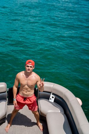 Shirtless person in red swim trunks and a backward cap flashing a peace sign while standing on a pontoon boat over turquoise lake water, sunny summer boating vibe