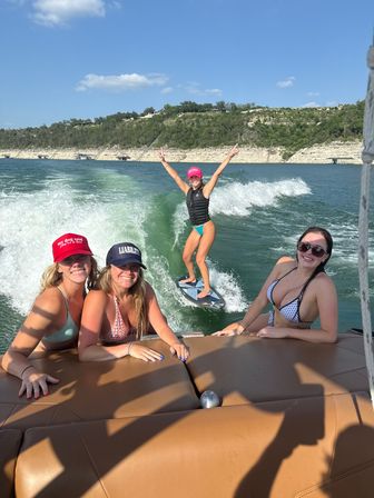 Four friends on a boat enjoying summer wakesurfing on a sunny lake with cliff-lined shoreline — one rider balancing on the wake with arms raised.