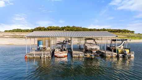 Sunny lake scene with a covered boat dock and multiple slips holding a wakeboard boat, a covered speedboat, and a pontoon with a slide, calm water and sandy shoreline with trees