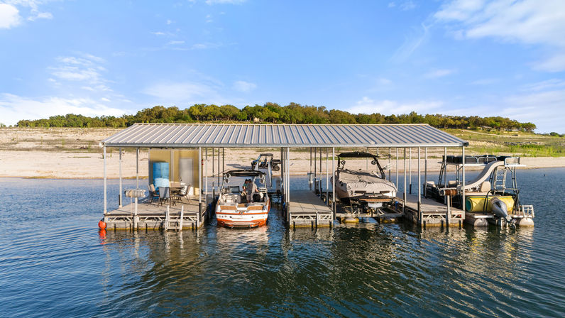 Sunny lake scene with a covered boat dock and multiple slips holding a wakeboard boat, a covered speedboat, and a pontoon with a slide, calm water and sandy shoreline with trees